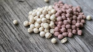 Deux tas de pois secs côte à côte sur une planche de bois gris : à gauche des graines nues de couleur naturelle, à droite les mêmes graines enrobées d'un rose crayeux de traitement