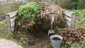 Tas de compost dans un bac de palettes, un creux creusé à la bêche montrant la matière qui fonce et s'affine vers le fond, bêche plantée, seau galvanisé et réserve de feuilles mortes à côté